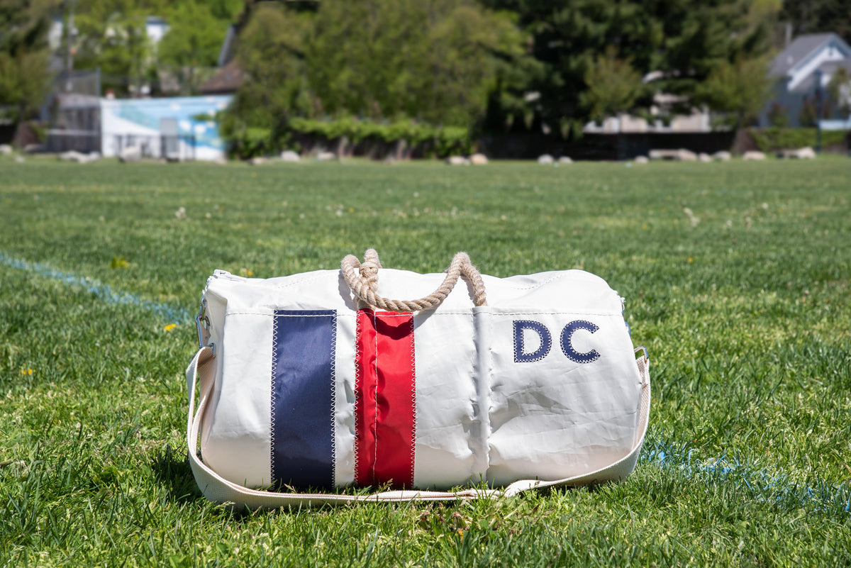 A white duffel bag with blue and red vertical stripes and the letters DC on it sits on green grass, with trees and buildings visible in the blurred background.