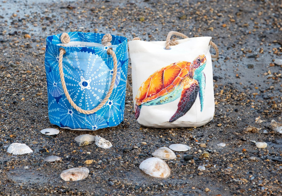 urchins bucket bag and watercolor sea turtle bucket bag perched on a pebbly beach