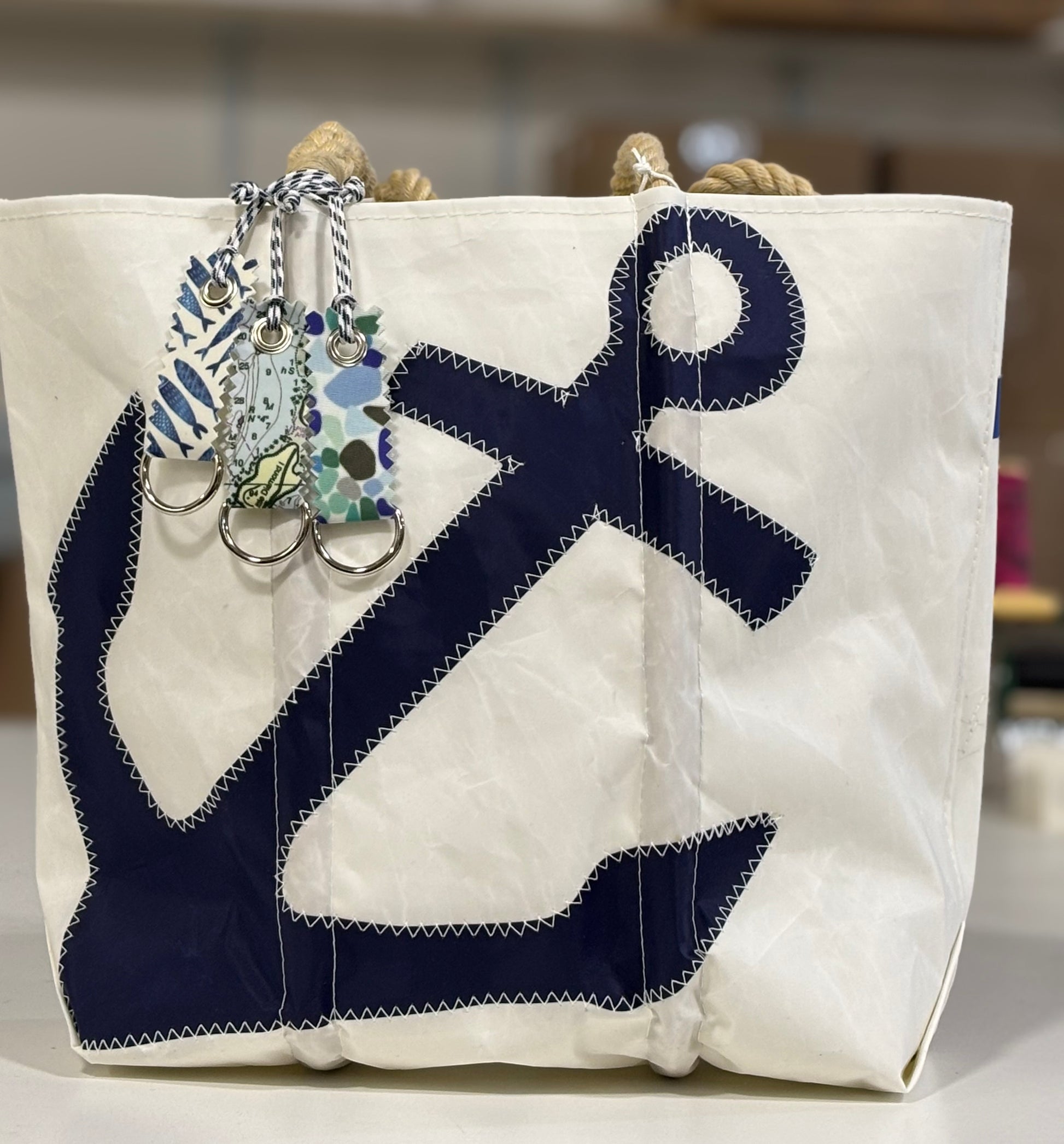 A white canvas tote with rope handles and a bold navy anchor design is topped with three patterned fabric keychains—including the Sea Bags Fish Print Sail Bag Charm—for an effortlessly sustainable look. Accented with upcycled sail cloth, this bag offers eco-conscious fashion with unmistakable nautical flair. by Sea Bags