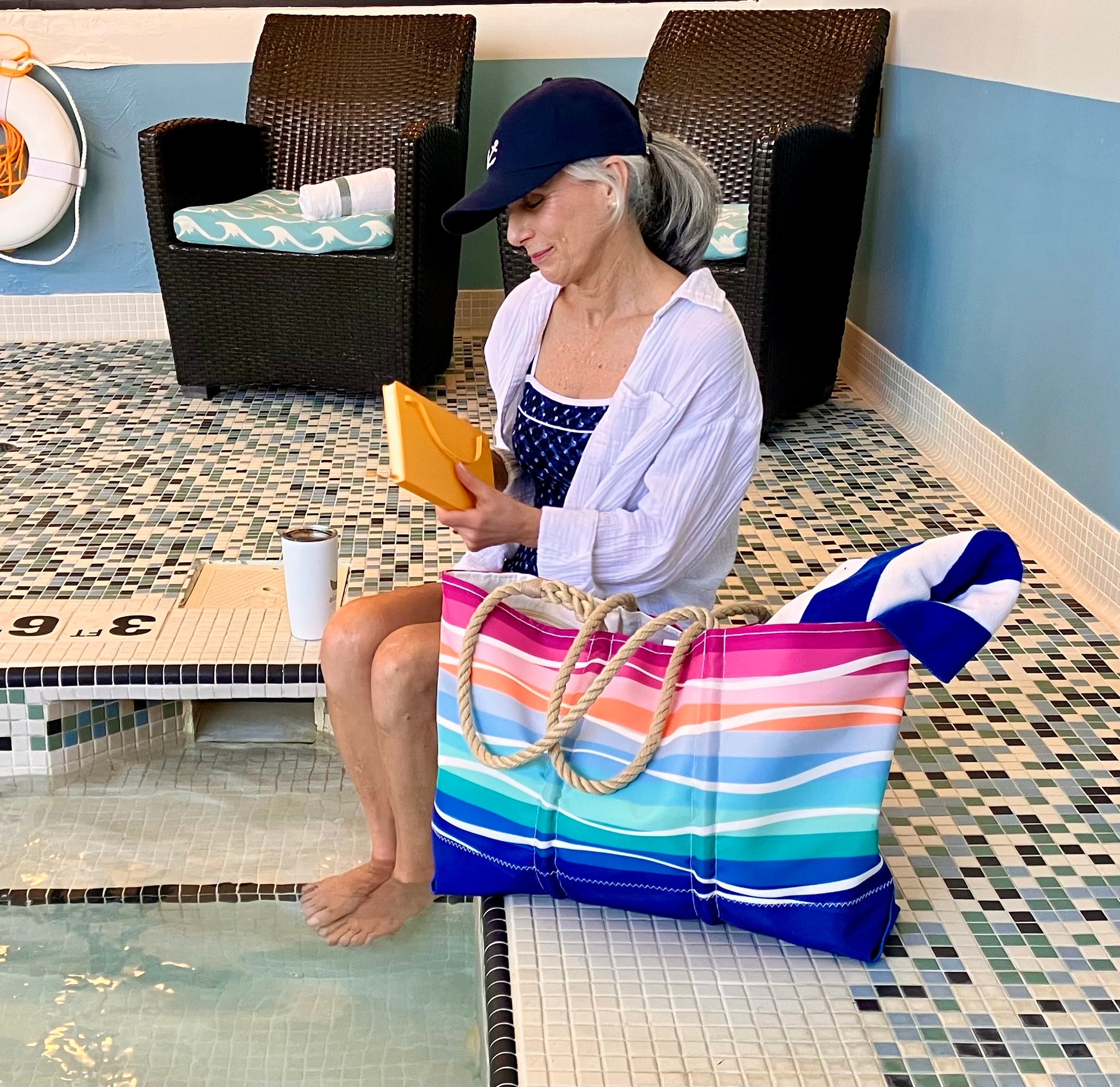 An older woman sits by an indoor pool with her feet in the water, holding a book, while the Sea Bags Sunset Surf Ogunquit Beach Tote—with its colorful design, sturdy rope handles, and waterproof pockets—rests beside her. by Sea Bags