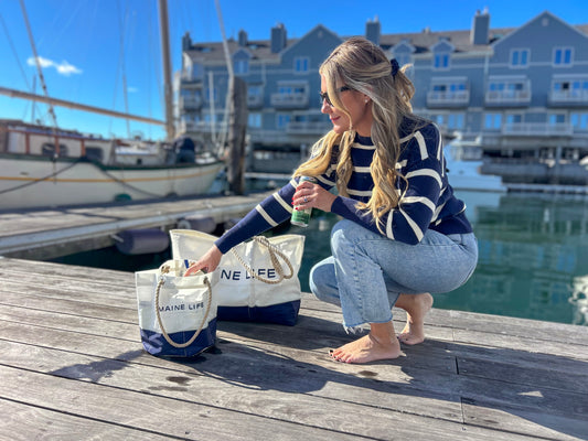 A woman in a striped sweater and jeans crouches barefoot on a dock, reaching into the Maine Life Beverage Bucket by Sea Bags—crafted from recycled sail cloth—with boats and buildings in the background. by Sea Bags