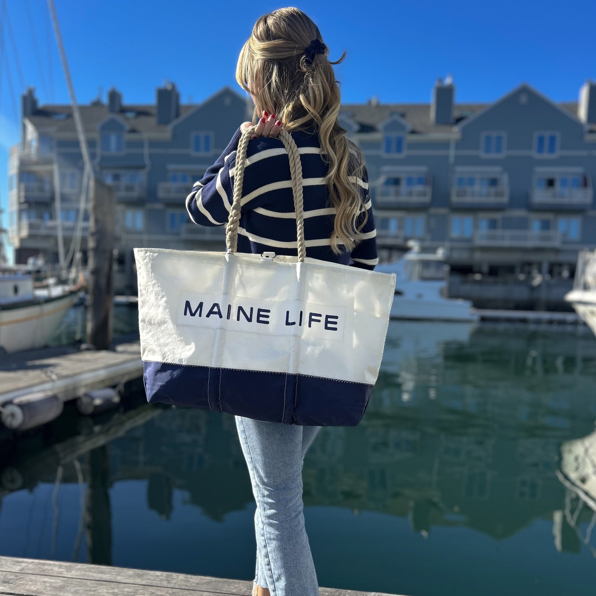 A woman with long, wavy hair holds the Maine Life Large Tote by Sea Bags near a marina, boats and waterfront buildings in the background on a sunny day—an effortless portrait that captures the spirit of Maine Life as curated by Erin Ovalle. by Sea Bags