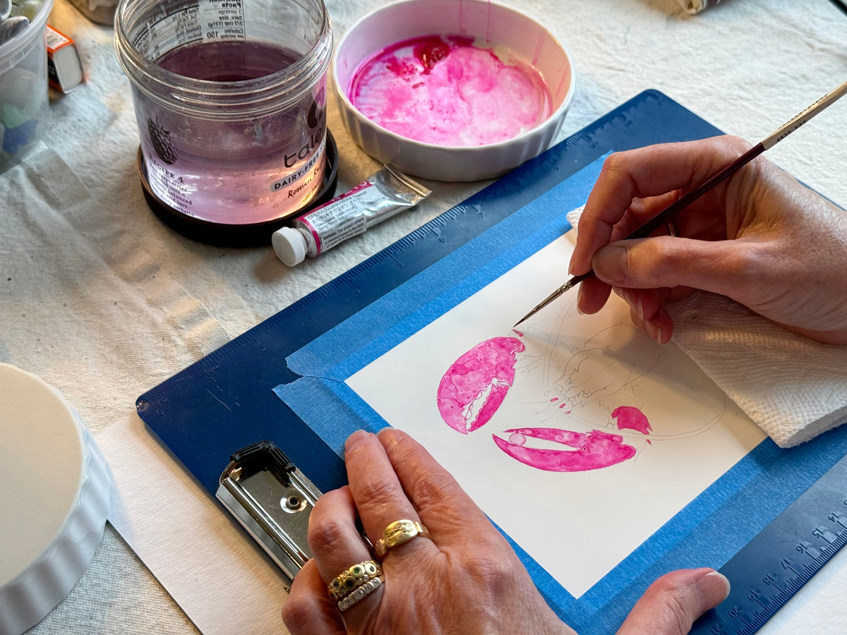 A Maine artist uses a fine paintbrush to paint the Pink Lobster design, inspired by a lobster, in watercolor on white paper, with painting supplies, a paint jar, and a palette nearby on a blue clipboard—the artwork featured on the Sea Bags Pink Lobster Medium Tote. by Sea Bags