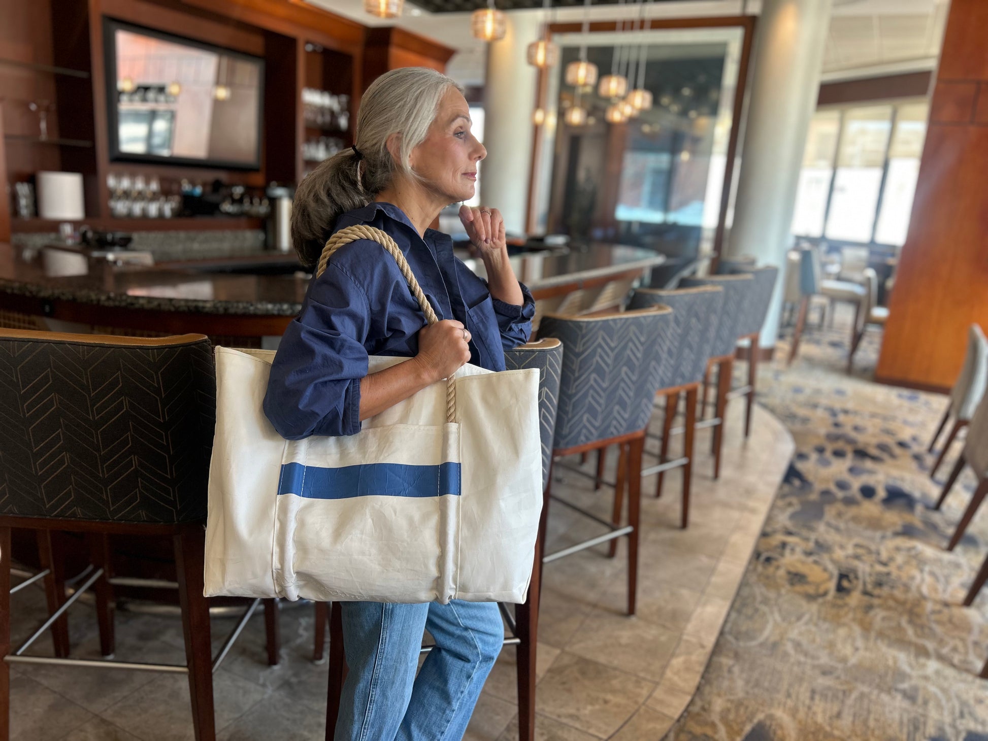 A woman with gray hair stands in a bar, dressed in a navy jacket and jeans, holding the Sea Bags Trolley Sleeve Travel Tote—a large white bag crafted from recycled sailcloth with blue stripes, rope handles, and a convenient mesh trolley sleeve. by Sea Bags