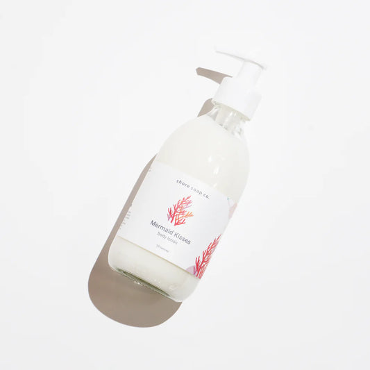 A clear pump bottle filled with white liquid labeled Mermaid Kisses Eco-Friendly Body Lotion by Shore Soap Co rests on a white background, casting a soft shadow. This natural body lotion evokes the refreshing essence of the sea with every use. by Sea Bags
