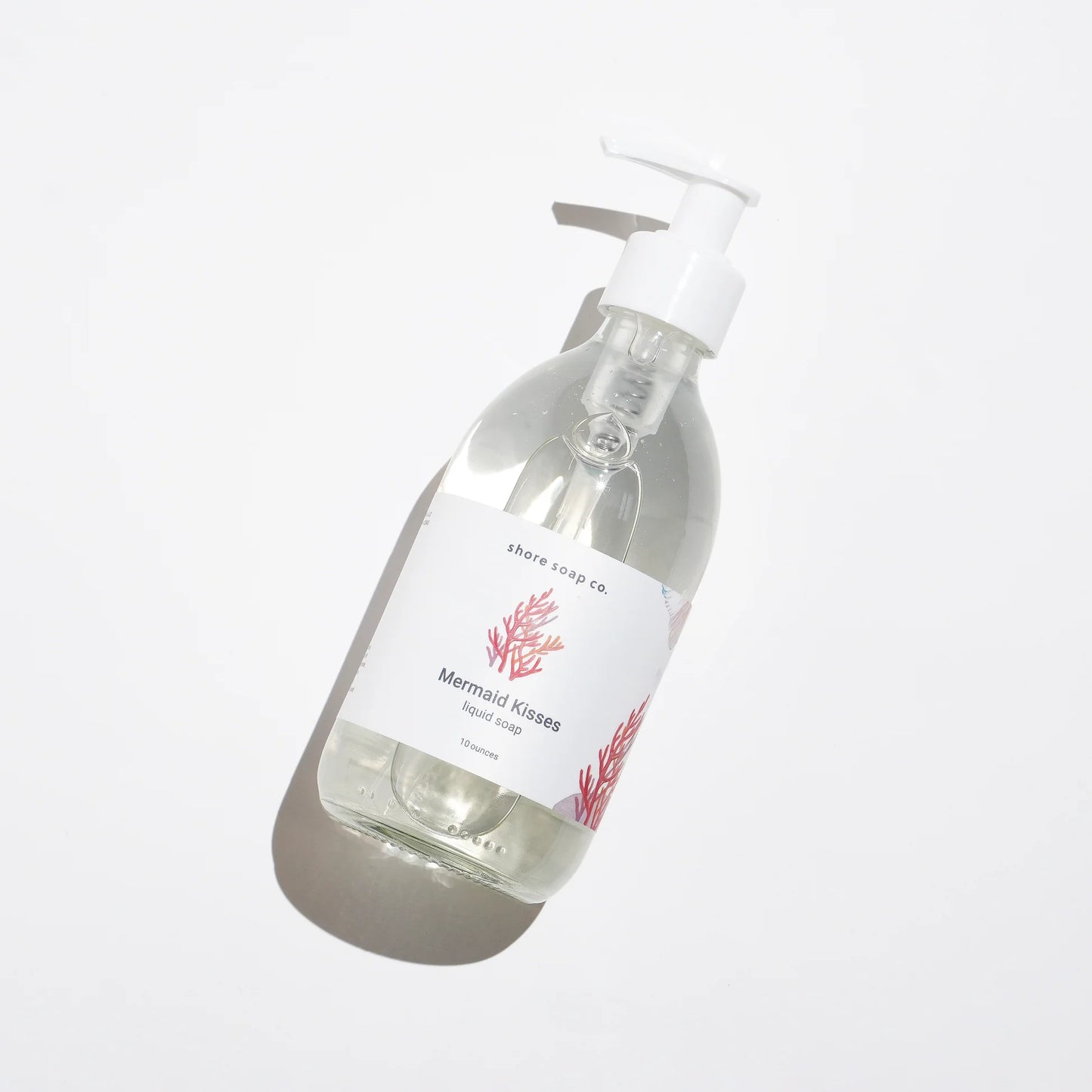 A clear pump bottle of Mermaid Kisses Eco-Friendly Liquid Soap by Shore Soap Co., adorned with a red coral graphic, rests on a white background and casts a soft shadow. Made with natural ingredients, this gentle hand soap is perfect for sensitive skin.