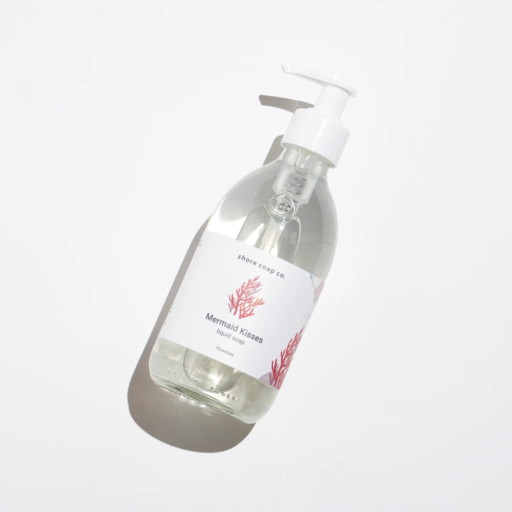 A clear pump bottle of Mermaid Kisses Eco-Friendly Liquid Soap by Shore Soap Co., adorned with a red coral graphic, rests on a white background and casts a soft shadow. Made with natural ingredients, this gentle hand soap is perfect for sensitive skin.