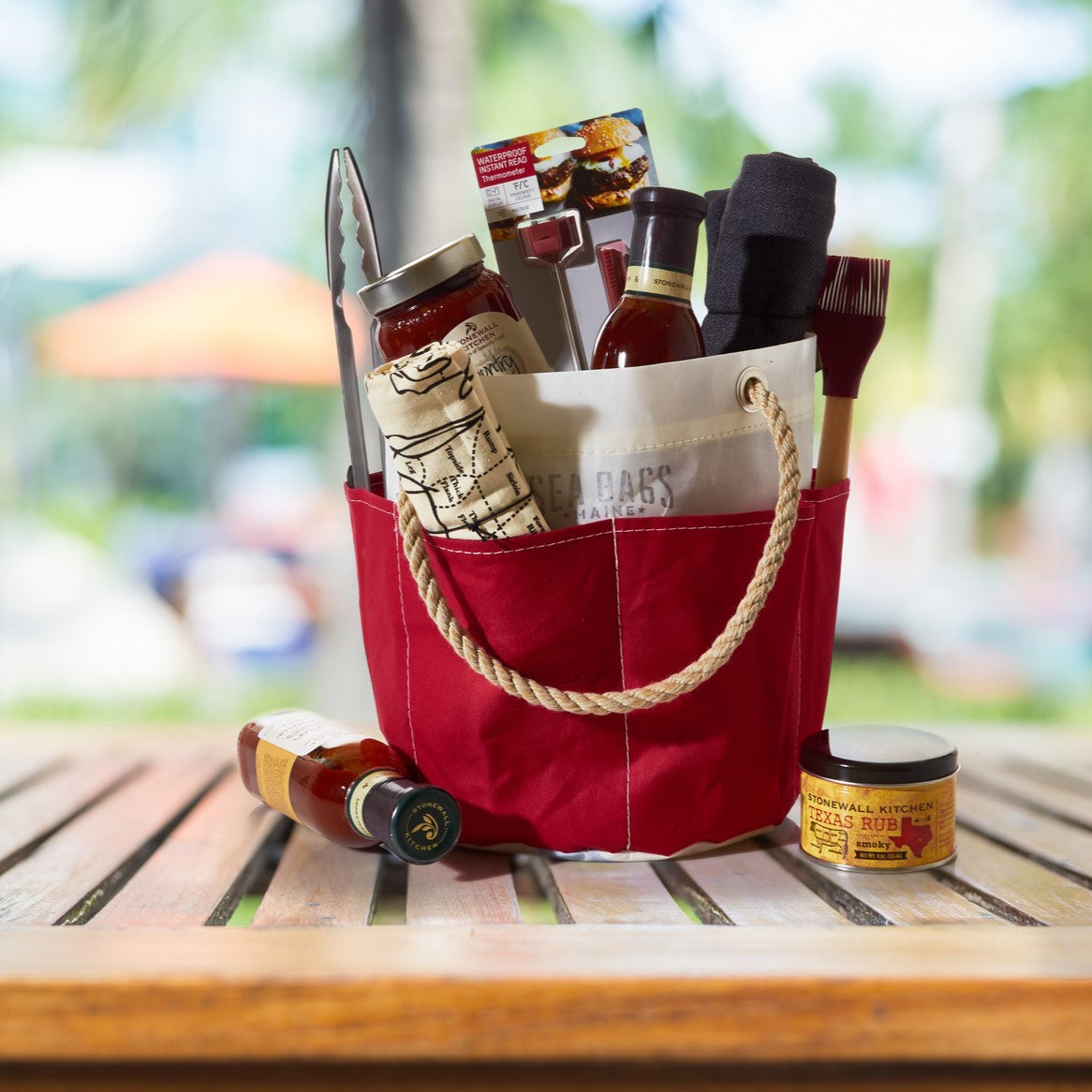 The All Purpose Red Tool Bucket Bag by Sea Bags, crafted from durable red canvas and equipped with handy exterior pockets for organization, sits outdoors on a wooden table. Its filled with barbecue tools, sauces, spice rub, and a cloth, standing out against a softly blurred background. by Sea Bags