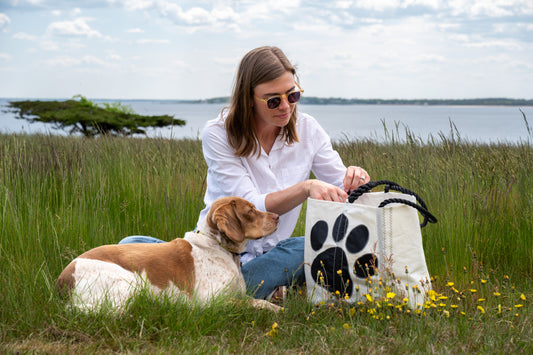 A woman in sunglasses sits on the grass by the water with her dog, reaching into her Sea Bags Paw Print Medium Tote with Clasp and Hanging Pocket—a white recycled sailcloth bag featuring a bold black paw print. Yellow flowers surround them. by Sea Bags
