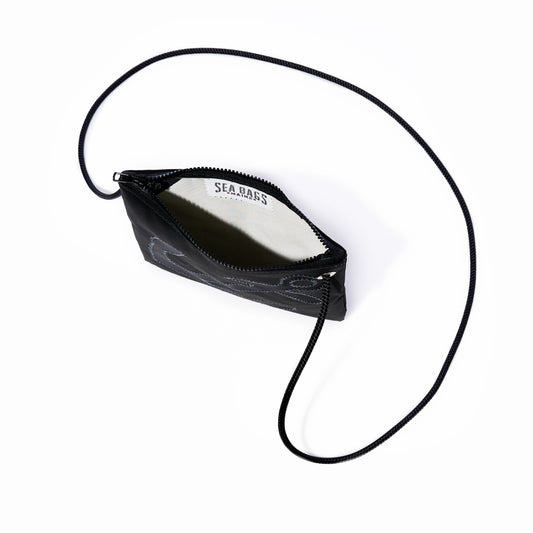 The Black-on-Black Anchor Slim Crossbody by Sea Bags is a small black zippered pouch featuring a thin, adjustable strap. Shown open to display its white interior label stamped with the Sea Bags brand, this hands-free bag is photographed against a white background. by Sea Bags