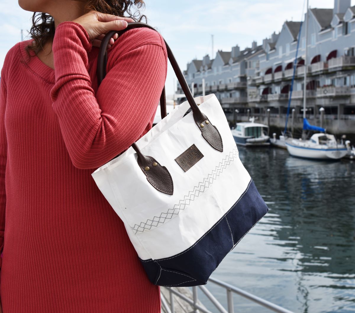 A person in a red dress holds the Chebeague Handbag by Sea Bags, a white and navy nautical bag with brown straps, standing by a marina with boats and buildings in the background. by Sea Bags