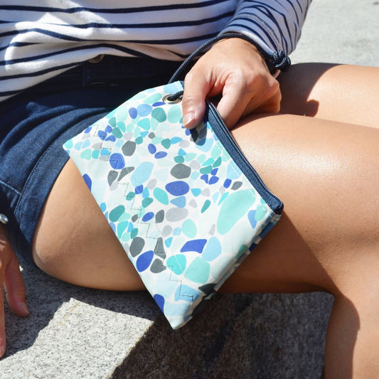 A person holds the Sea Bags Sea Glass Print Wristlet while sitting outdoors, dressed in a navy skirt and striped shirt. by Sea Bags