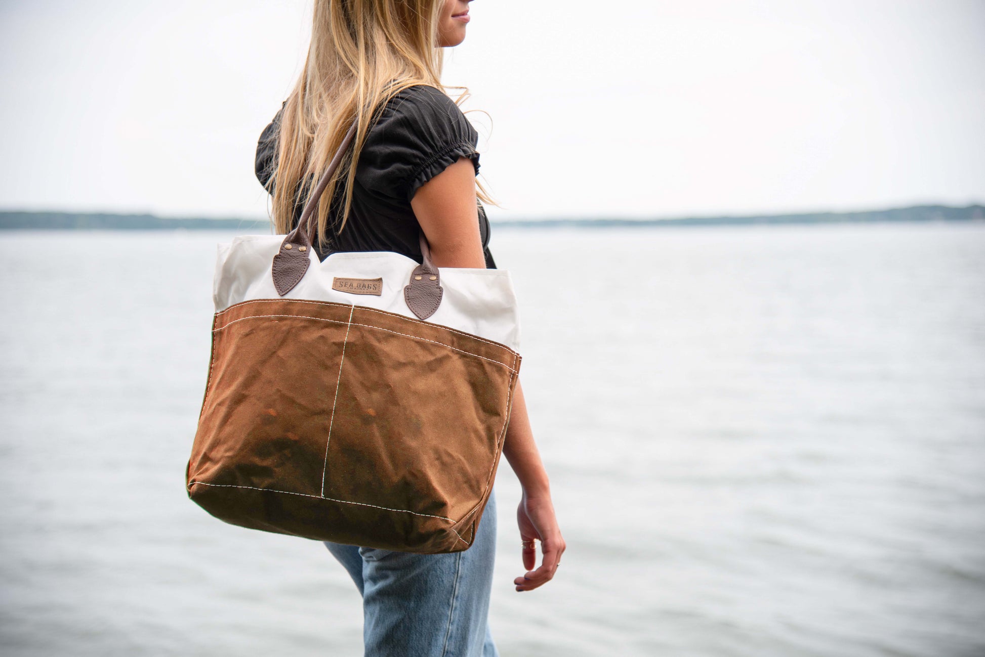A woman stands by the water holding a Sea Bags Tan Chebeague Tote, crafted from recycled sail cloth, while wearing a black shirt and jeans. by Sea Bags