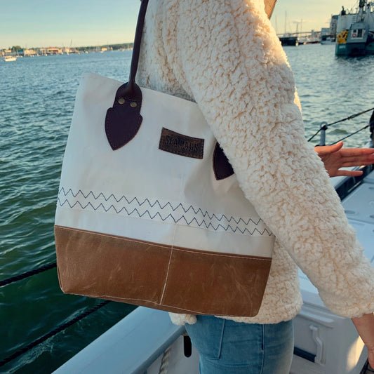 Wearing a cream fleece jacket and blue jeans, a person stands on a boat near a harbor holding the Sea Bags Tan Chebeague Handbag. by Sea Bags