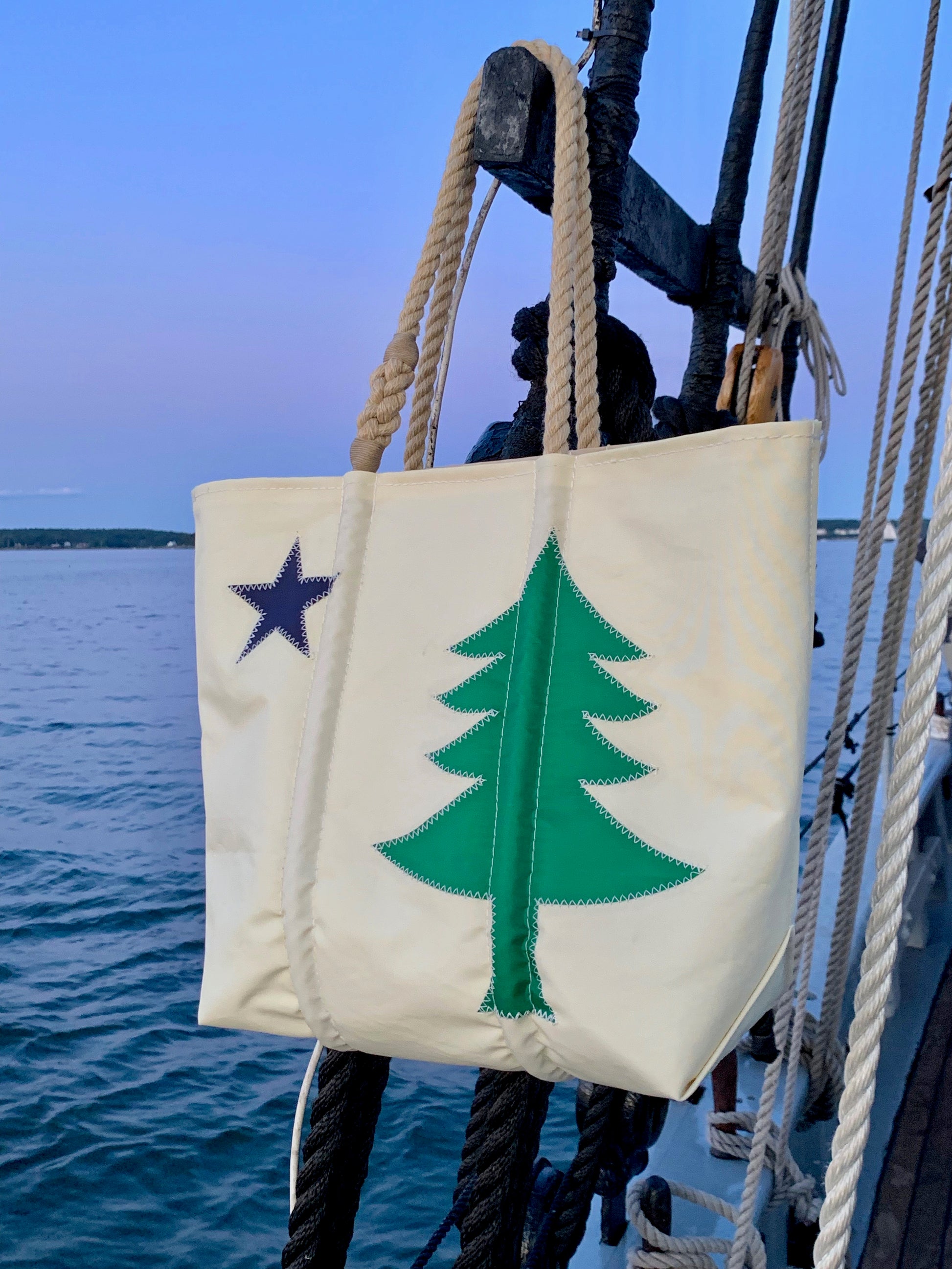 A Sea Bags Maine Bicentennial Medium Tote, crafted from recycled sail cloth with rope handles and adorned with a green pine tree and a blue star inspired by the Maine state flag, hangs on the railing of a boat at sunset. by Sea Bags