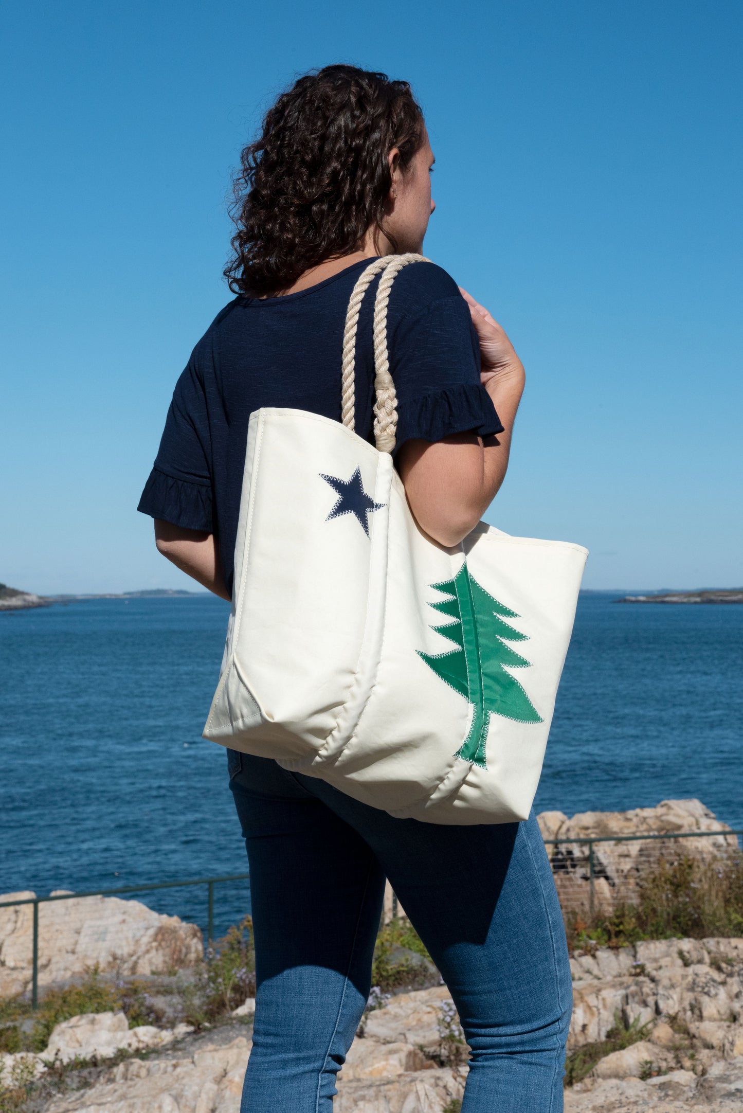 A person stands by the ocean holding the Sea Bags Maine Bicentennial Medium Tote, a white recycled sail cloth bag featuring a green tree and black star inspired by the Maine state flag. by Sea Bags