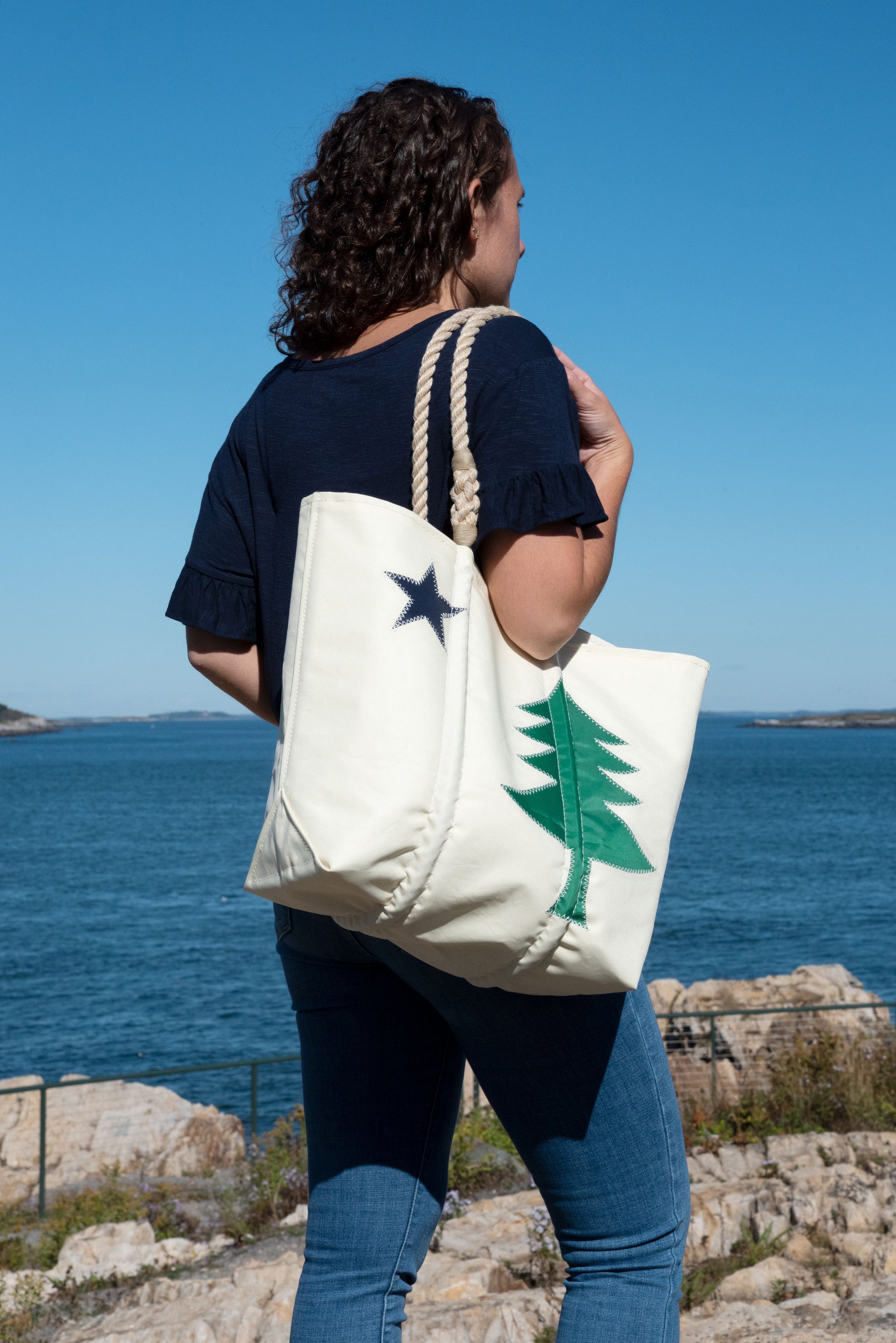 A person stands by the ocean holding the Sea Bags Maine Bicentennial Medium Tote, a white recycled sail cloth bag featuring a green tree and black star inspired by the Maine state flag. by Sea Bags