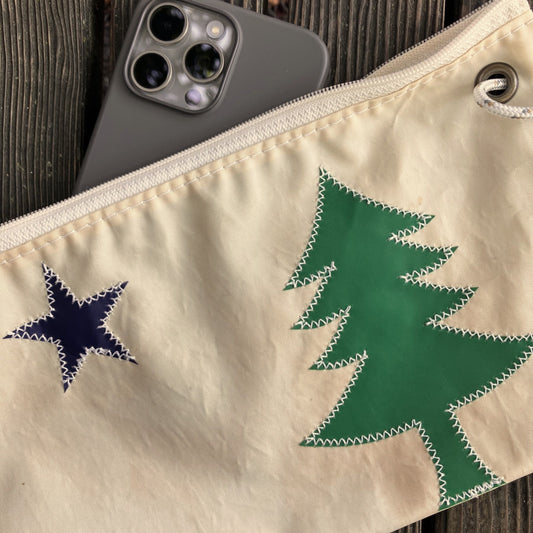 A Sea Bags Maine Bicentennial Large Wristlet, adorned with a blue star and a green tree reminiscent of the Maine state flag, sits on a wooden surface with a smartphone partially tucked inside its beige fabric. by Sea Bags