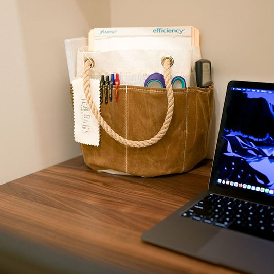 The Sea Bags All Purpose Tan Tool Bucket Bag, crafted from durable canvas with waxed-canvas pockets, neatly holds folders, pens, scissors, and a calculator on a wooden desk next to an open laptop. by Sea Bags