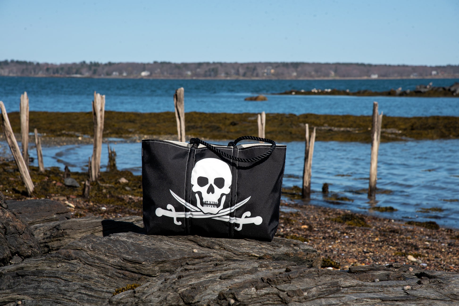 The Sea Bags Jolly Roger Tote with Clasp and Hanging Pocket, made from recycled sail cloth, showcases a bold white skull and crossed swords design on its black exterior. The tote is pictured resting on rocks by the shoreline, with the sea and a wooden fence in the background. by Sea Bags