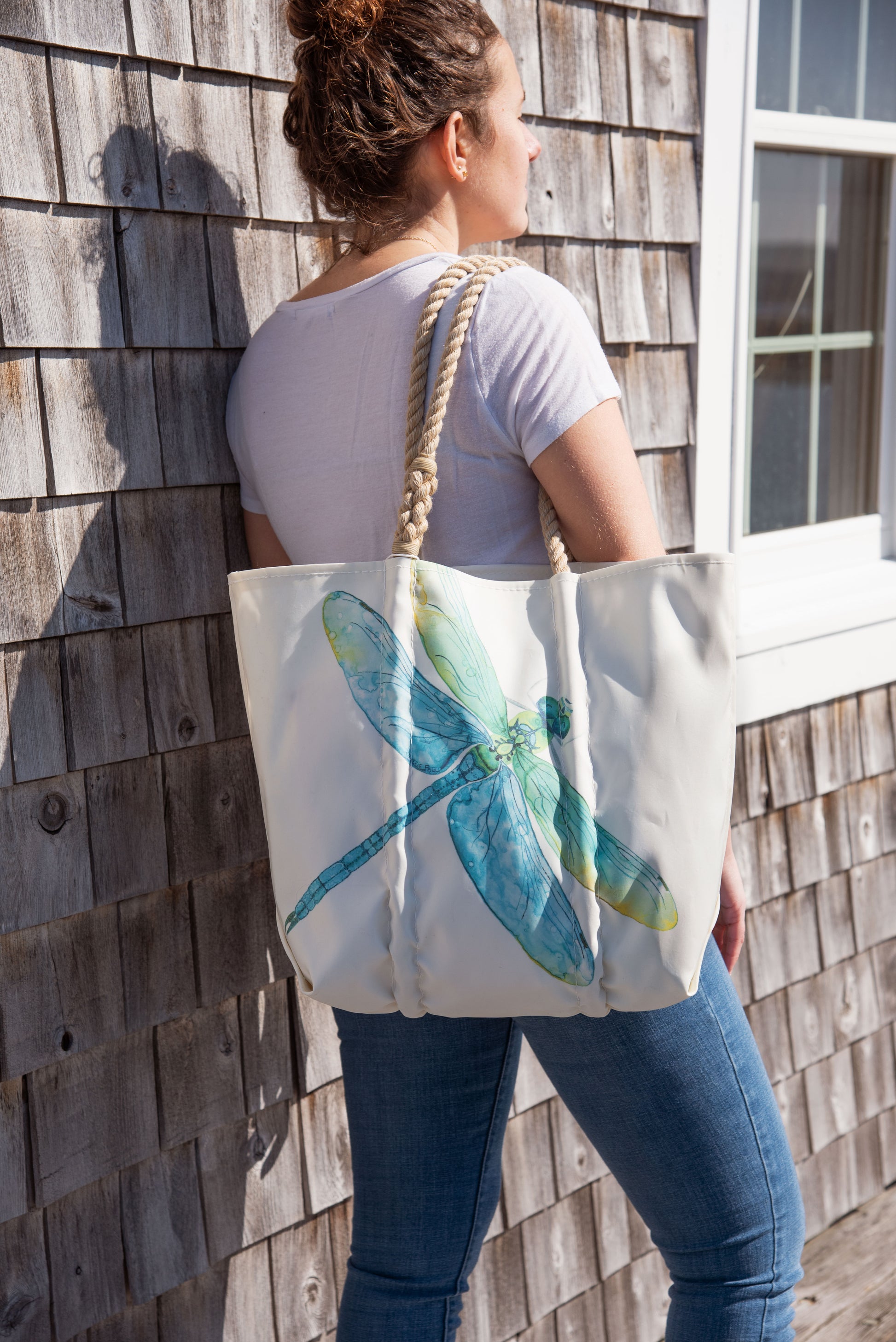 A woman in a white t-shirt and jeans stands outside by a shingled wall, carrying the Sea Bags Watercolor Dragonfly Medium Tote with thick rope handles, featuring an original design created by a local artist. by Sea Bags