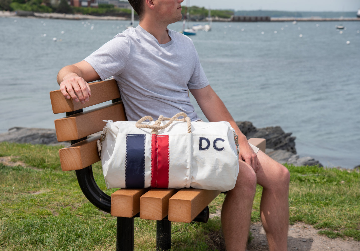 A person in a gray shirt sits on a wooden bench by the water with a Sea Bags Custom Monogram Vertical Stripe Duffel resting beside them. by Sea Bags