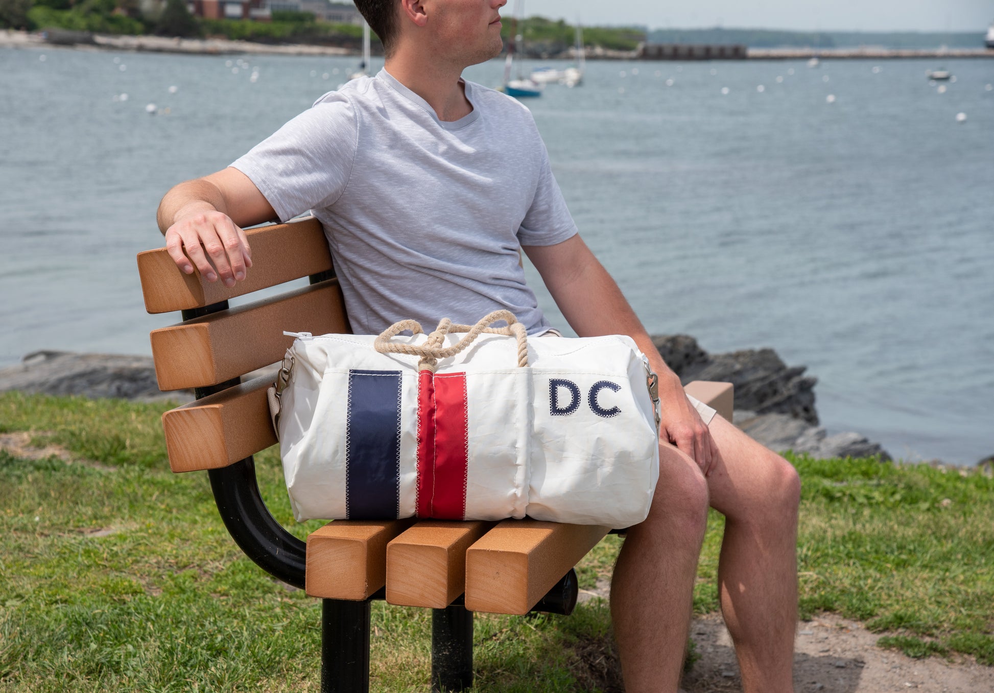 A person in a gray shirt sits on a wooden bench by the water with a Sea Bags Custom Monogram Vertical Stripe Duffel resting beside them. by Sea Bags