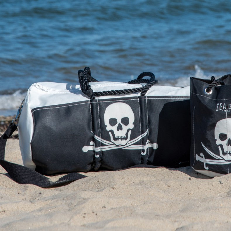 The Sea Bags Jolly Roger Duffel, expertly crafted from recycled sails and featuring a striking skull and crossbones design in black and white, sits on the sand near the ocean next to a matching tote.