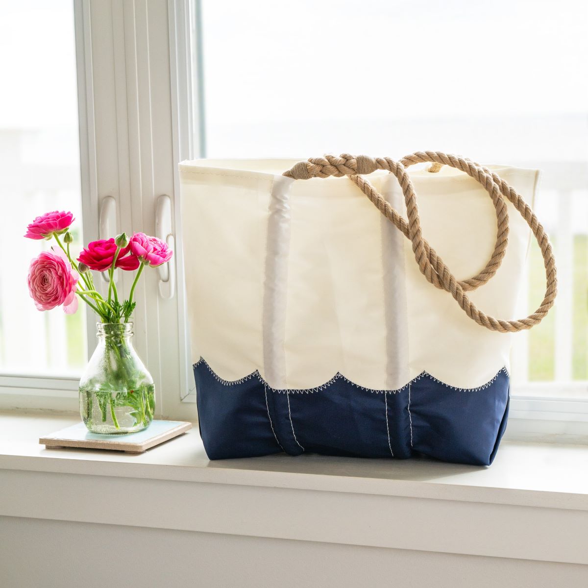 The Wavy Seas Medium Tote by Sea Bags, crafted from recycled sail cloth and featuring blue and white nautical stripes with sturdy rope handles, sits on a windowsill beside a glass vase of pink flowers. by Sea Bags