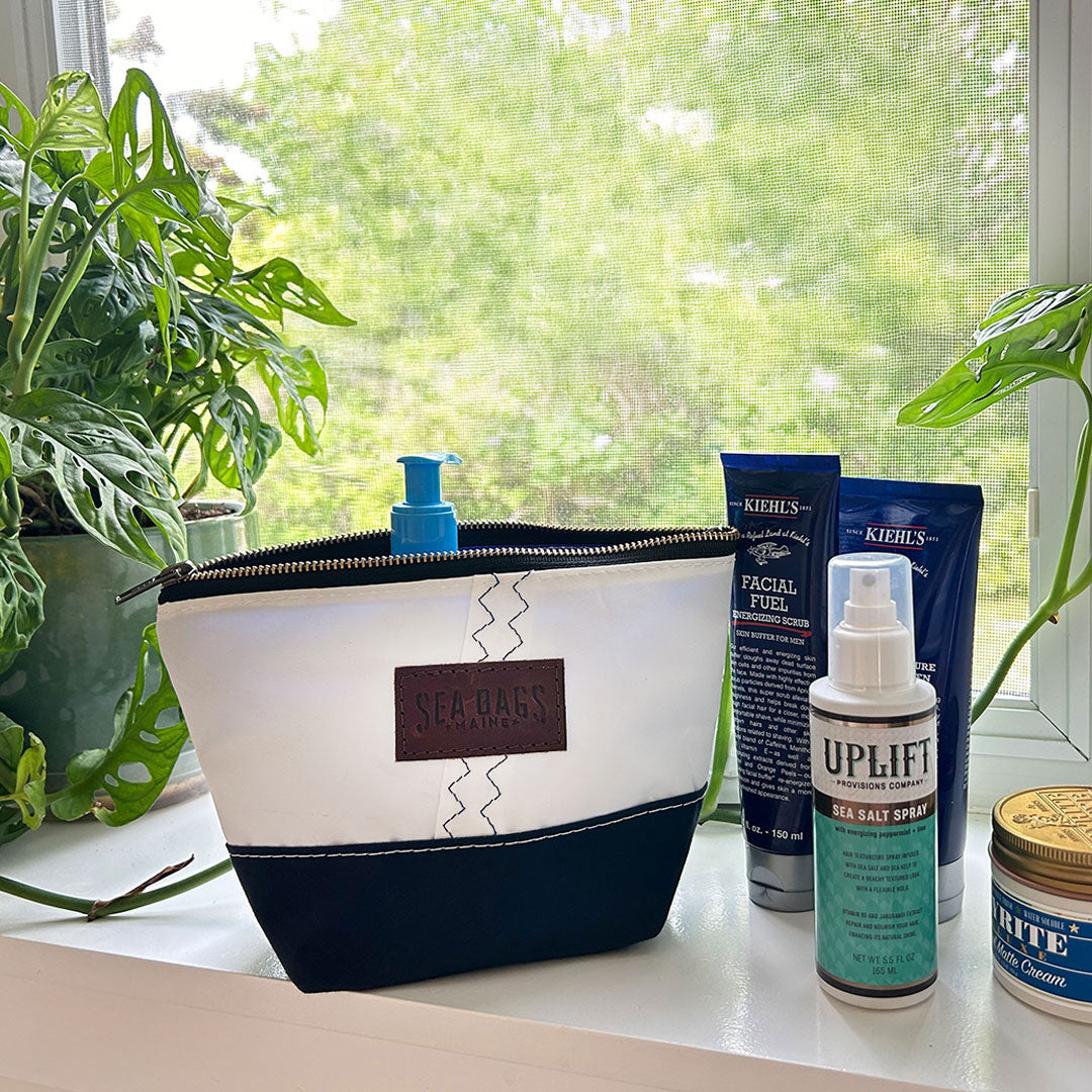The Sea Bags Chebeague Cosmetic Bag, crafted from canvas and leather, is arranged among various grooming products on a windowsill, with green plants and natural light in the background. by Sea Bags