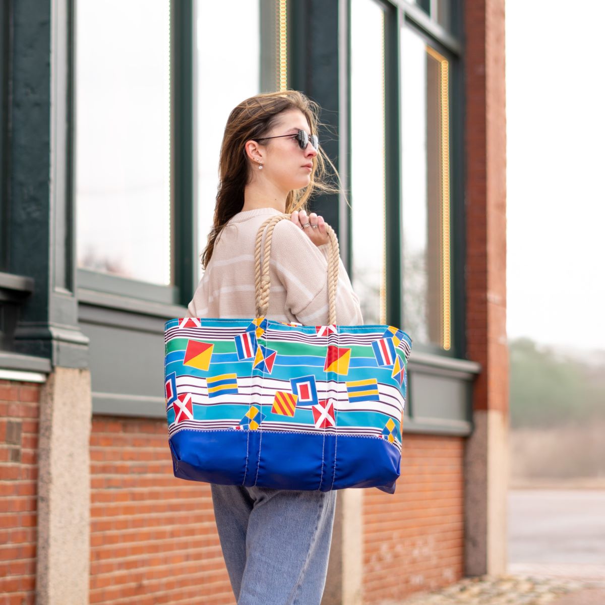 A woman wearing sunglasses stands outside a building, carrying the Sea Bags Signal Flags Ogunquit Beach Tote—crafted from recycled sail cloth and featuring rope handles—over her shoulder. by Sea Bags