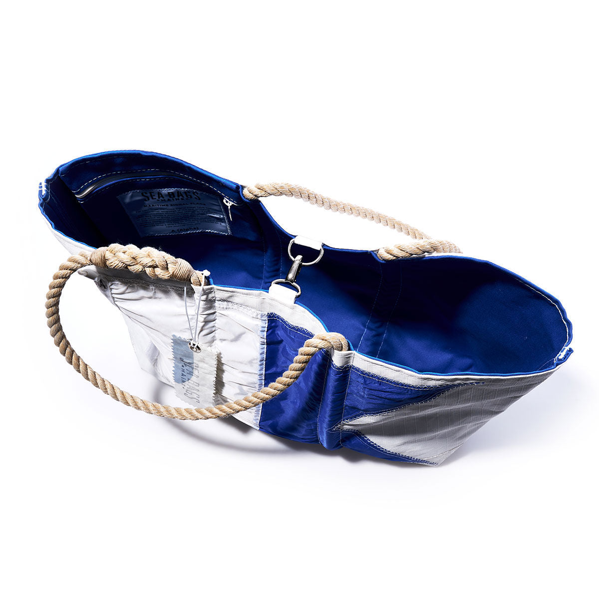 Sea Bags Alpha Signal Flag Large Tote features a blue and silver design crafted from recycled sail cloth, rope handles, an open top revealing a blue interior, metal clasp closure, and an attached tag inspired by vintage signal flags. by Sea Bags