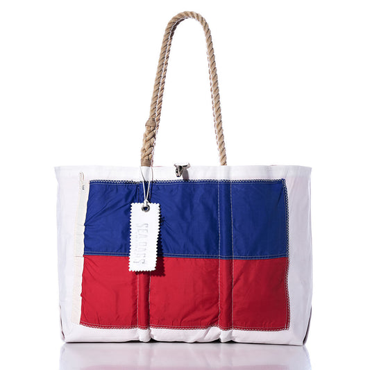 The Echo Signal Flag Large Tote by Sea Bags is a white tote bag with rope handles, crafted from recycled sail cloth. Its front panel is divided horizontally with blue on top and red on the bottom, drawing inspiration from vintage signal flags for a nautical flair. A tag hangs from one handle, adding to the stylish design. by Sea Bags