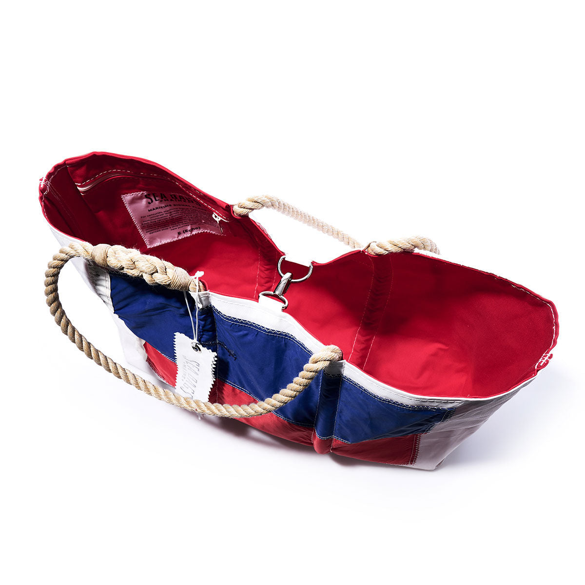 The Sea Bags Echo Signal Flag Large Tote features an open design with sturdy rope handles, a vibrant red interior, and a nautical-themed exterior in blue, white, and red inspired by vintage signal flags. Crafted from recycled sail cloth, this tote includes a white tag attached to one handle. by Sea Bags