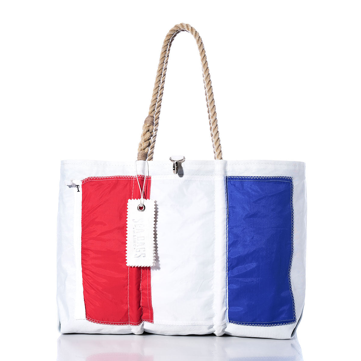 Introducing the Sea Bags Tango Signal Flag Large Tote—a spacious white tote bag crafted from recycled sail cloth, featuring bold red, white, and blue vertical panels inspired by vintage maritime signal flags. Detailed with sturdy rope handles and a signature white tag attached to the handle, this tote blends nautical charm with sustainable style. by Sea Bags