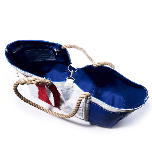 The Sea Bags Tango Signal Flag Large Tote, crafted from recycled sail cloth and inspired by maritime signal flags, features bold blue, white, and red panels, sturdy rope handles, and a blue interior lining. Shown from above with its top open, this bag blends nautical heritage with sustainable style. by Sea Bags