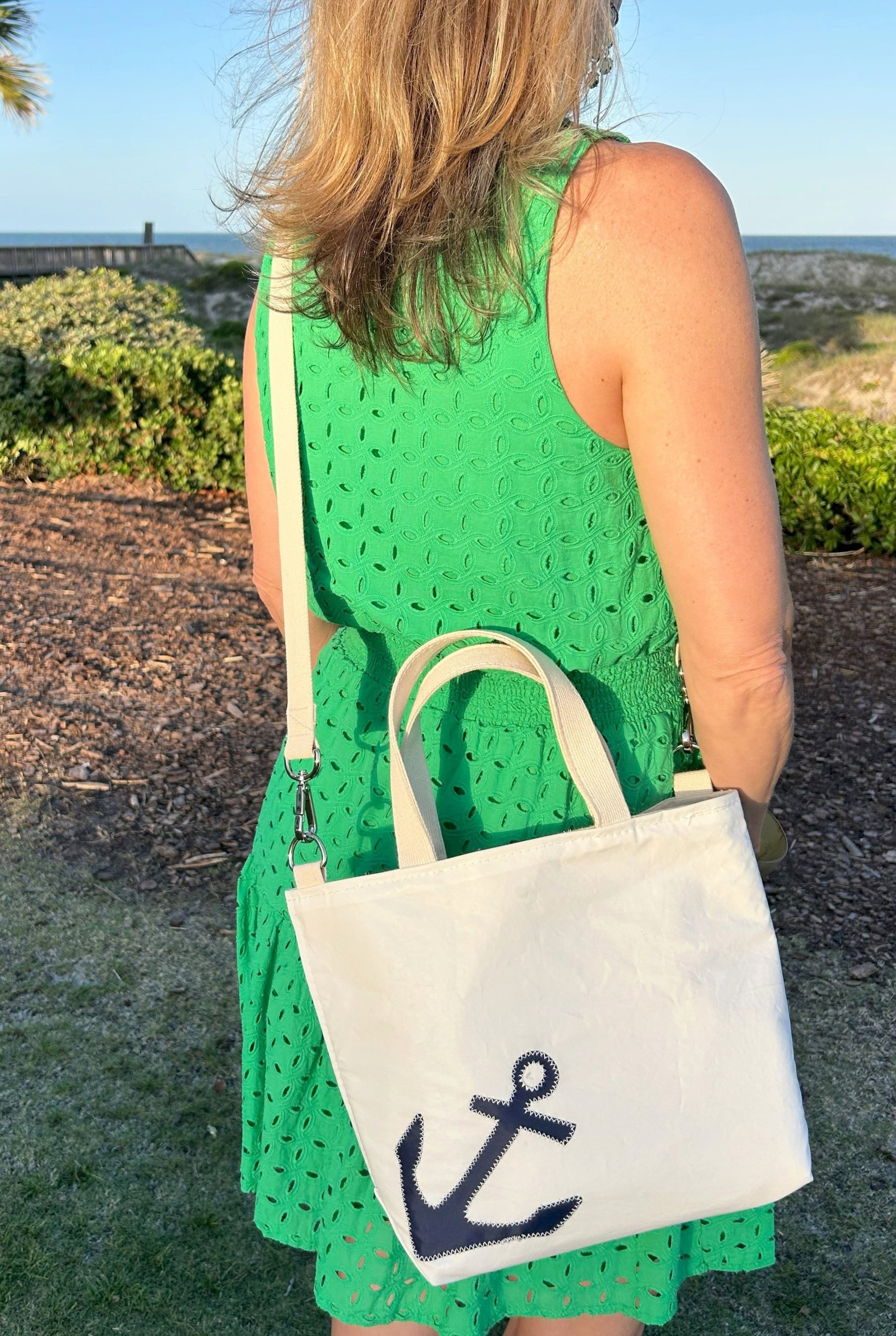 A woman in a green sleeveless dress stands outdoors, carrying the Sea Bags Navy Anchor Crossbody Tote—an eco-friendly bag made from recycled sailcloth and featuring a dark blue anchor design. by Sea Bags