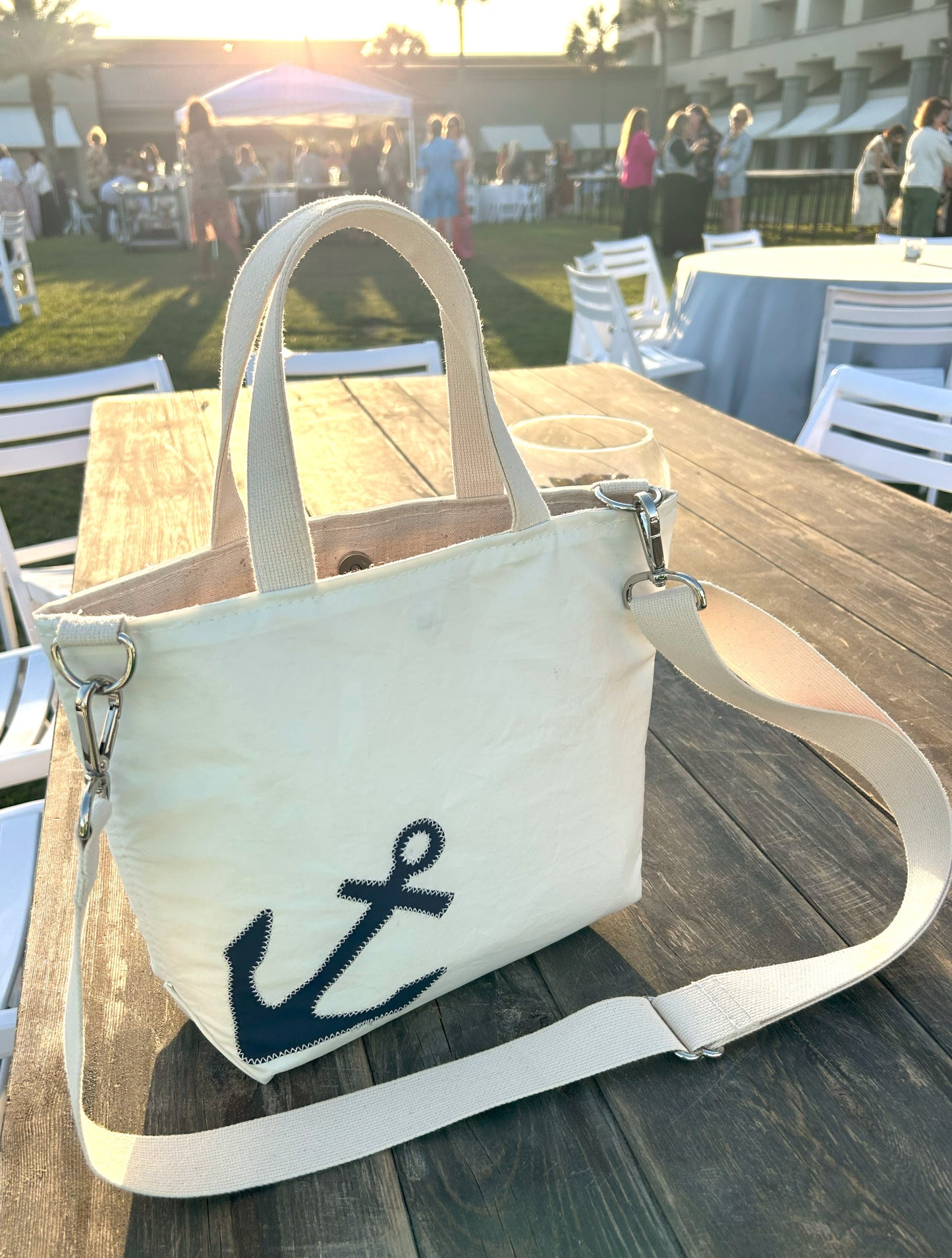 The Sea Bags Navy Anchor Crossbody Tote, an eco-friendly bag crafted from recycled sailcloth and featuring a navy anchor design, sits on a wooden table outdoors at a gathering with people and white chairs in the background. by Sea Bags