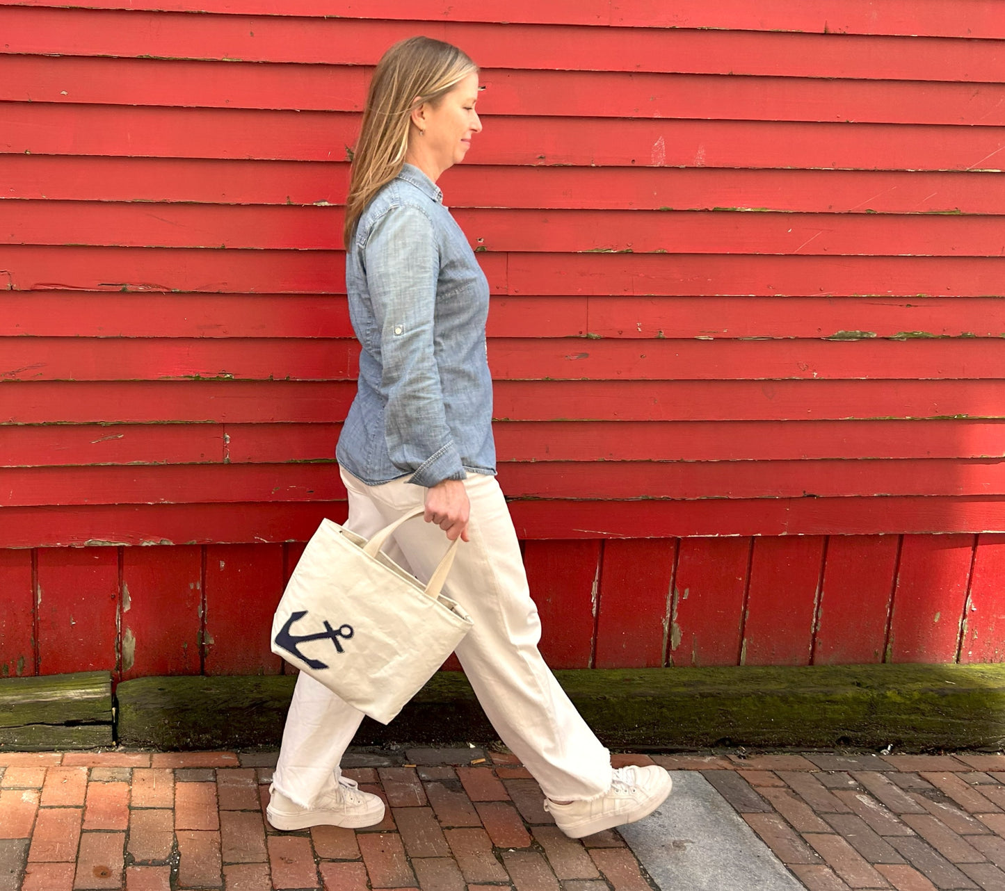 A woman walks on a brick sidewalk beside a red wooden wall, carrying the Sea Bags Navy Anchor Crossbody Tote—an eco-friendly bag crafted from recycled sailcloth and featuring a navy blue anchor design. by Sea Bags