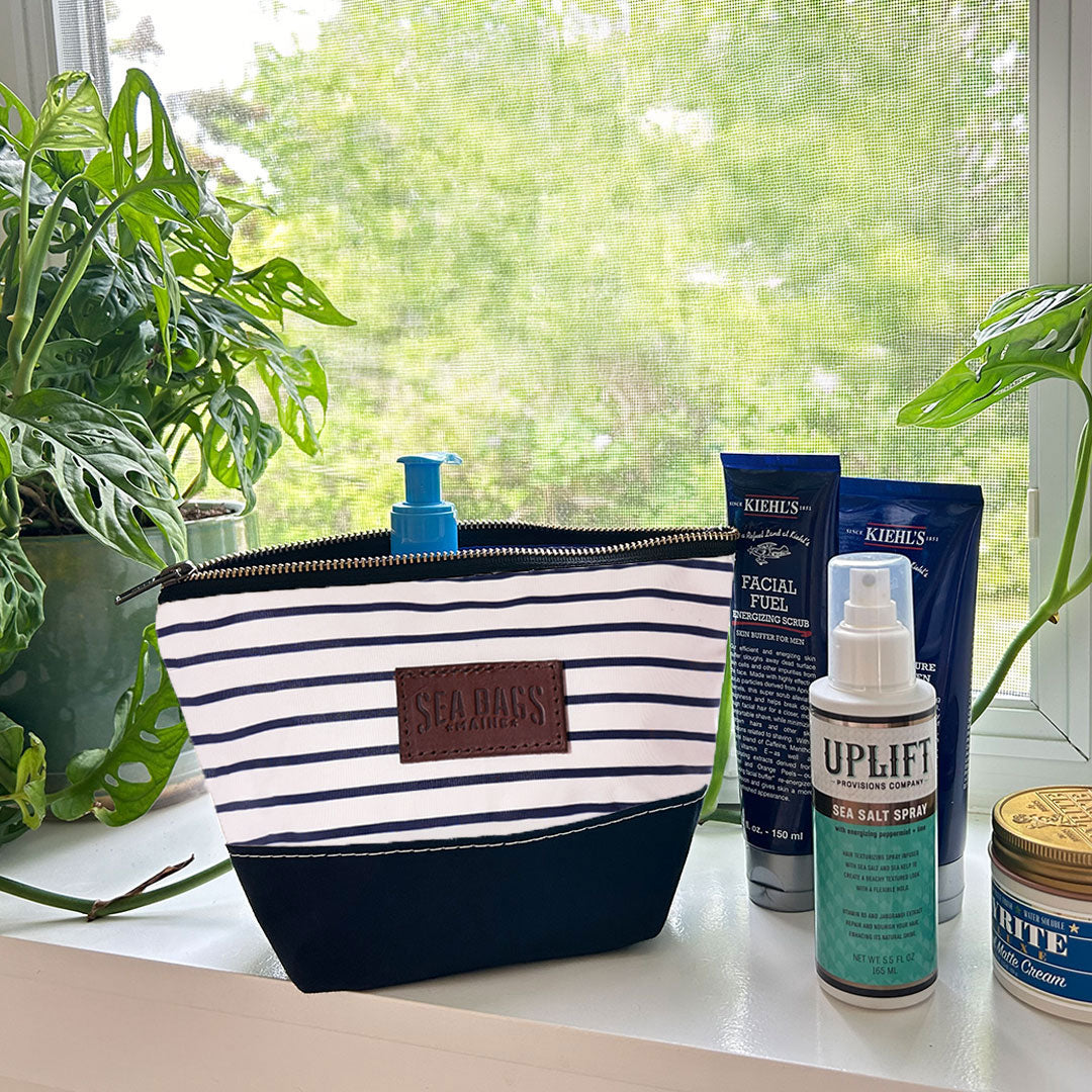 A Navy Mariner Stripe Chebeague Cosmetic Bag by Sea Bags, crafted from canvas and leather, sits on a windowsill beside skincare products, a can, and potted plants, with a lush green view outside the window. by Sea Bags