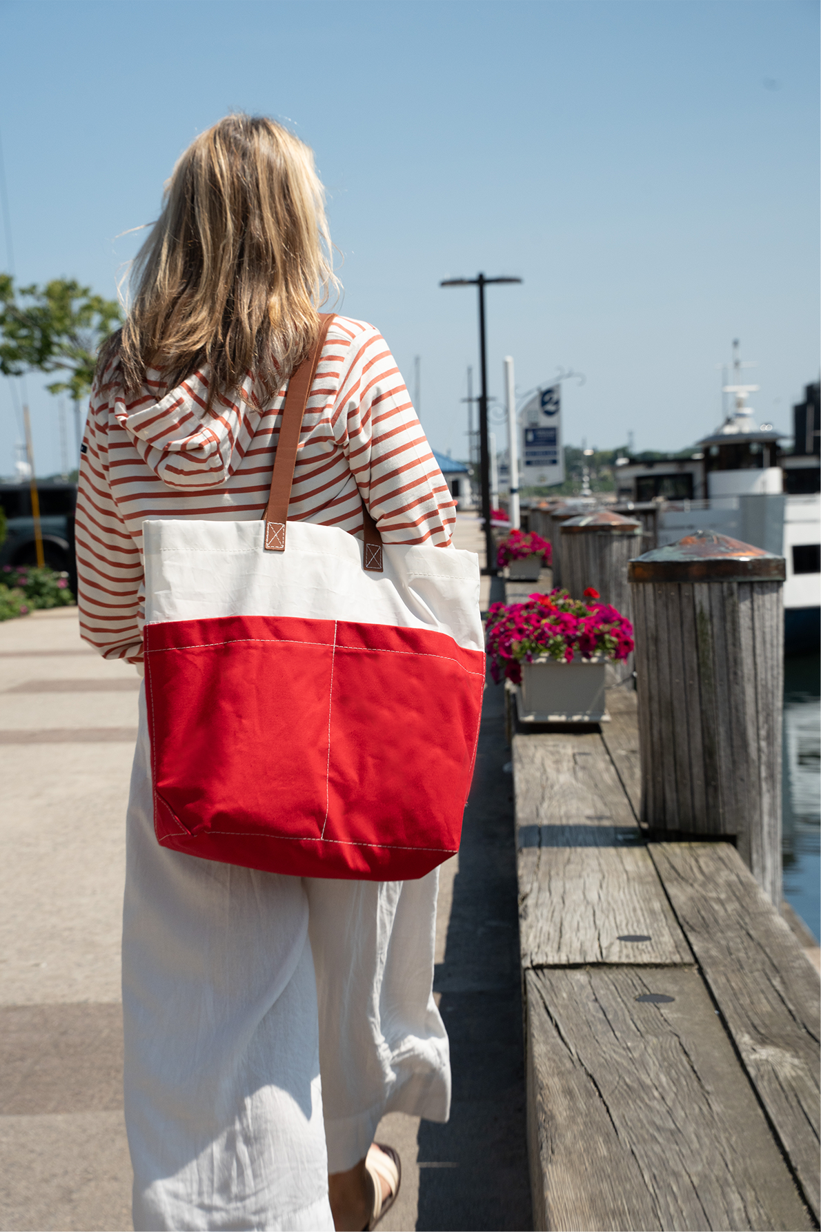 A person with blonde hair walks on a sunny waterfront path, wearing a striped hoodie and white pants, carrying a Red Chebeague Market Tote by Sea Bags, crafted from recycled sails with sturdy leather handles. by Sea Bags