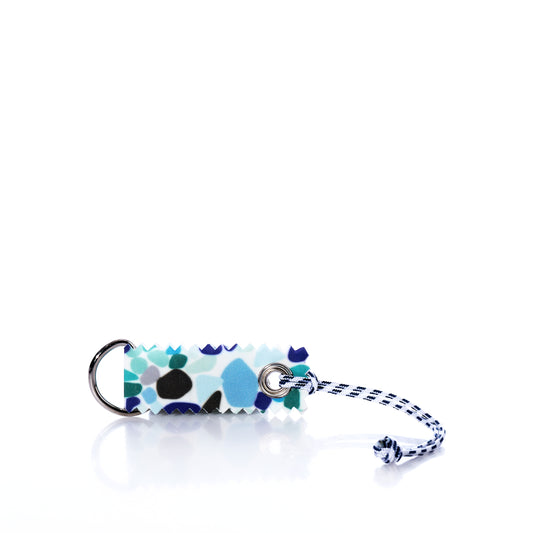 Sea Bags’ Sea Glass Print Sail Bag Charm features a rectangular design with a blue, black, and white abstract print, crafted from upcycled sail cloth for sustainable style. It is attached to a metal ring and finished with a blue-and-white cord, shown on a white background. by Sea Bags