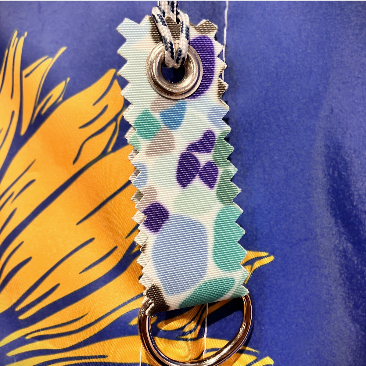 Close-up of the Sea Bags Sea Glass Print Sail Bag Charm, a vibrant fabric keychain crafted from upcycled sail cloth with distinctive zigzag edges, sturdy grommet, and metal ring—hanging against a blue and yellow background—for a sustainable and stylish addition to your collection. by Sea Bags