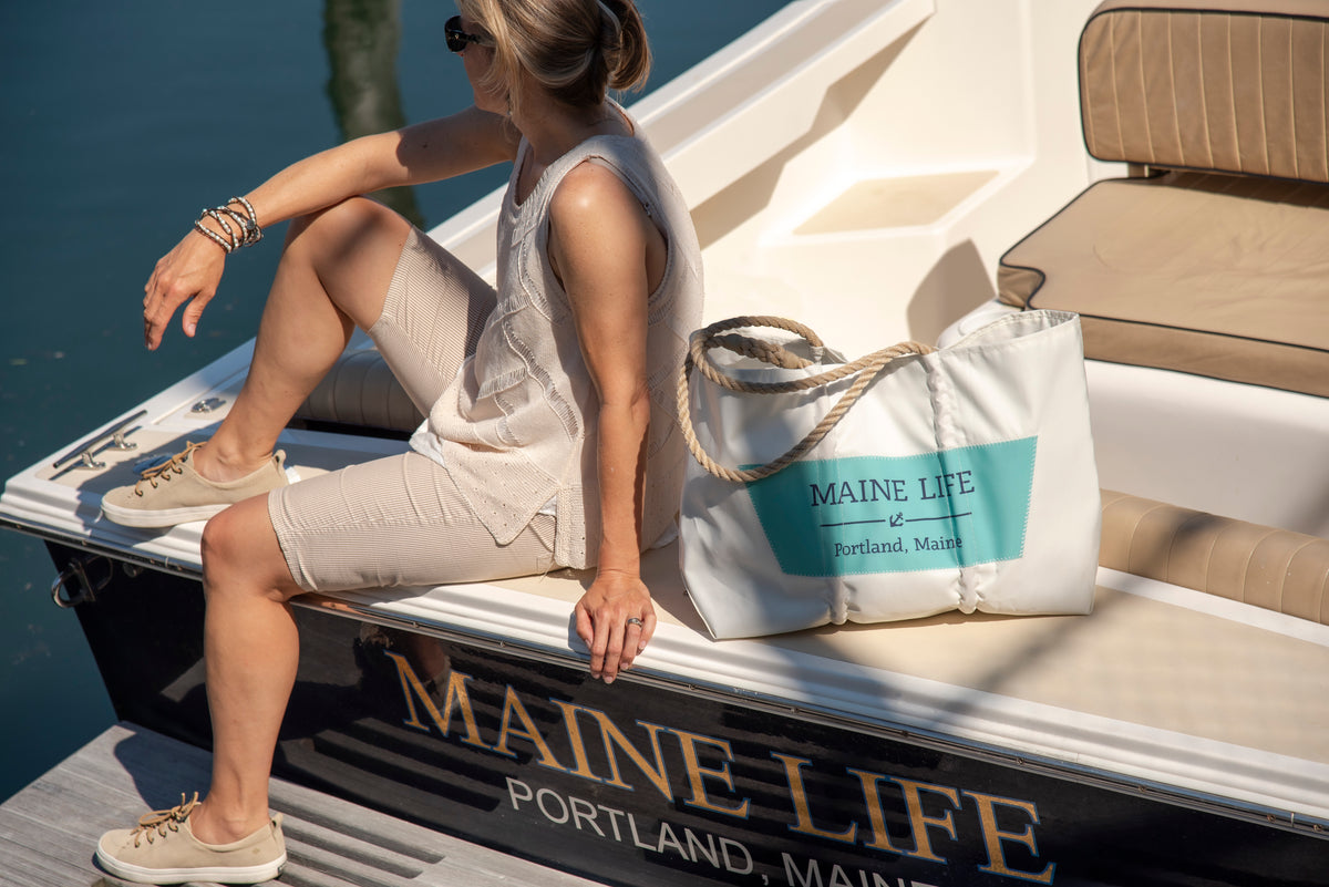 A woman in light clothing sits on a boat docked in Portland, Maine, with a large white Sea Bags Custom Boat Name Tote labeled Maine Life beside her. by Sea Bags