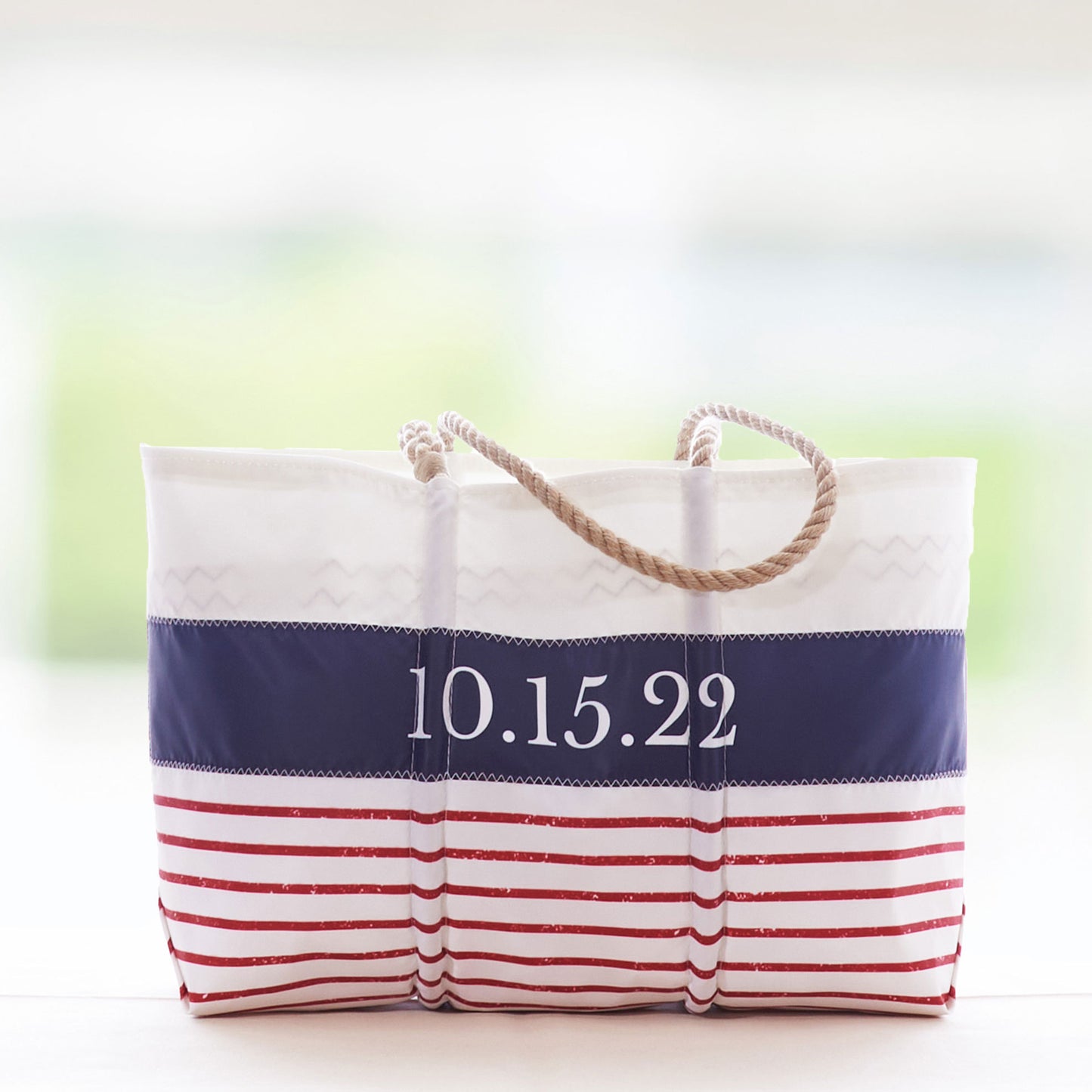The Sea Bags Custom Mariner Stripe Tote features a nautical design with red stripes, a blue stripe, and sturdy rope handles. Crafted from recycled sail cloth for style and sustainability, this white tote can be personalized with the date 10.15.22 printed on the front. by Sea Bags