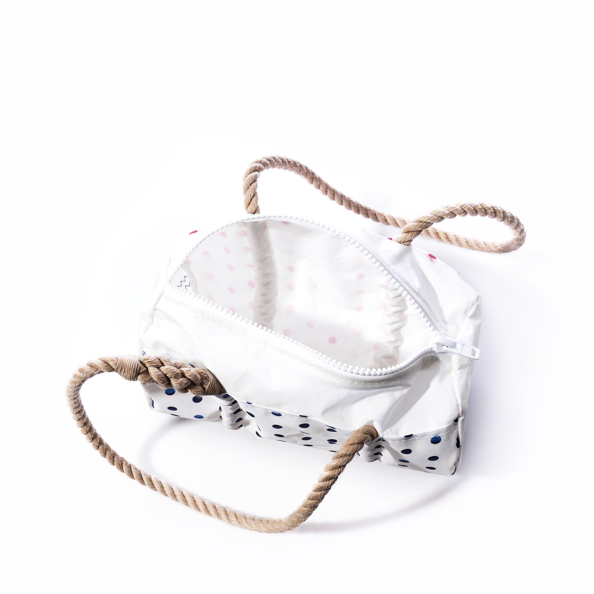 The Flip Side Summer Dots Baguette Bag by Sea Bags is an open, white rectangular zippered bag adorned with navy blue polka dots and features two thick beige rope handles, evoking the feel of a classic summer handbag, set against a plain white background. by Sea Bags