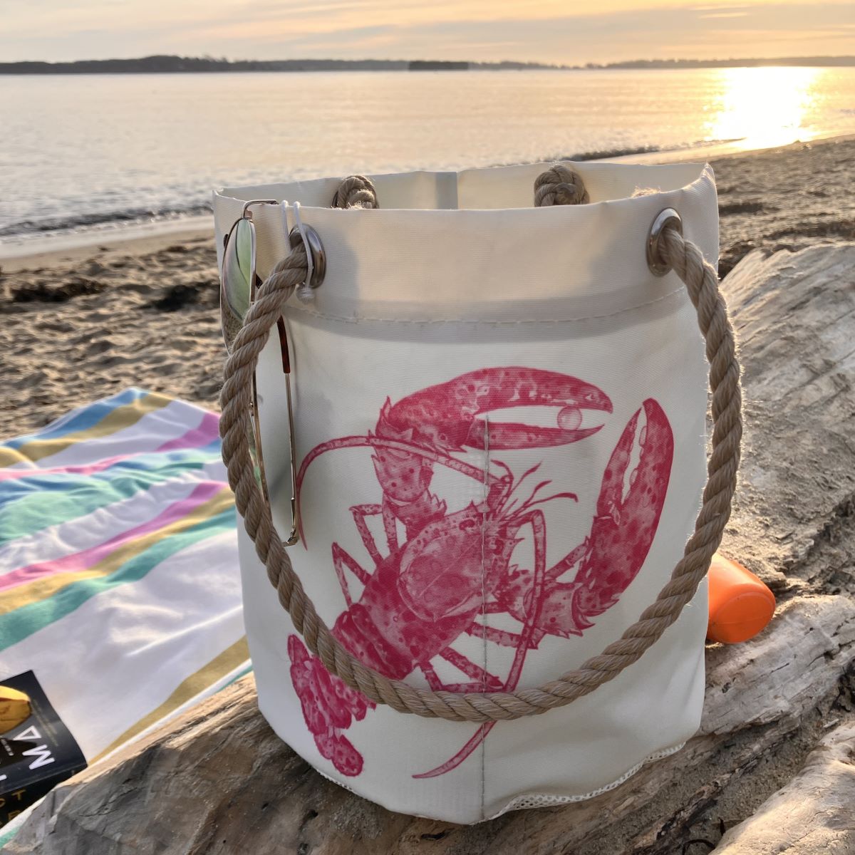 The Sea Bags Pink Lobster Beachcomber Bucket, made from recycled sail cloth and featuring pink lobster prints with sturdy rope handles, rests on a driftwood log beside a towel and sunscreen, as the sun sets over a sandy beach and calm water. by Sea Bags