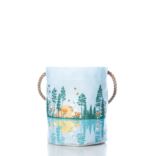 The Sea Bags Life is Good Lakeside Sunset Bucket is a charming light blue fabric storage basket with rope handles, adorned with a nature scene of trees, flowers, animals, and a yellow sun reflected in water—making it a delightful housewarming gift that helps keep your space beautifully organized. by Sea Bags