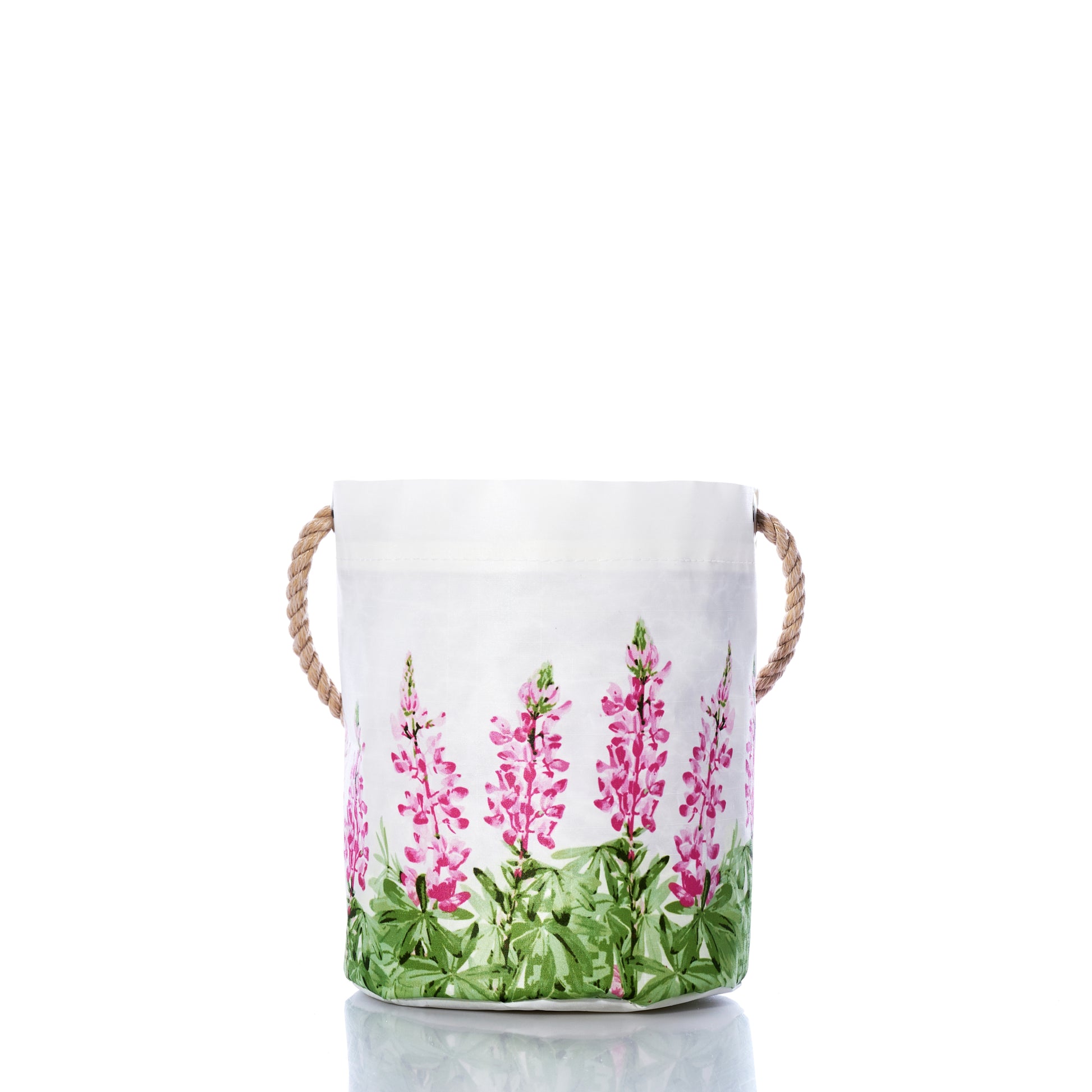 Sea Bags Pink Lupine Bucket features white fabric and sturdy rope handles, accented by a pink and green floral pattern near the base. Shown on a reflective surface with a white background, this stylish bucket adds a fresh touch of spring décor to any room. by Sea Bags