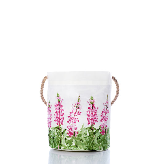 Sea Bags Pink Lupine Bucket—a white fabric storage basket with rope handles, adorned with a green leaf and pink flower design inspired by lupine fields—photographed on a white background with a subtle reflection at the base, making it perfect for fresh spring décor. by Sea Bags
